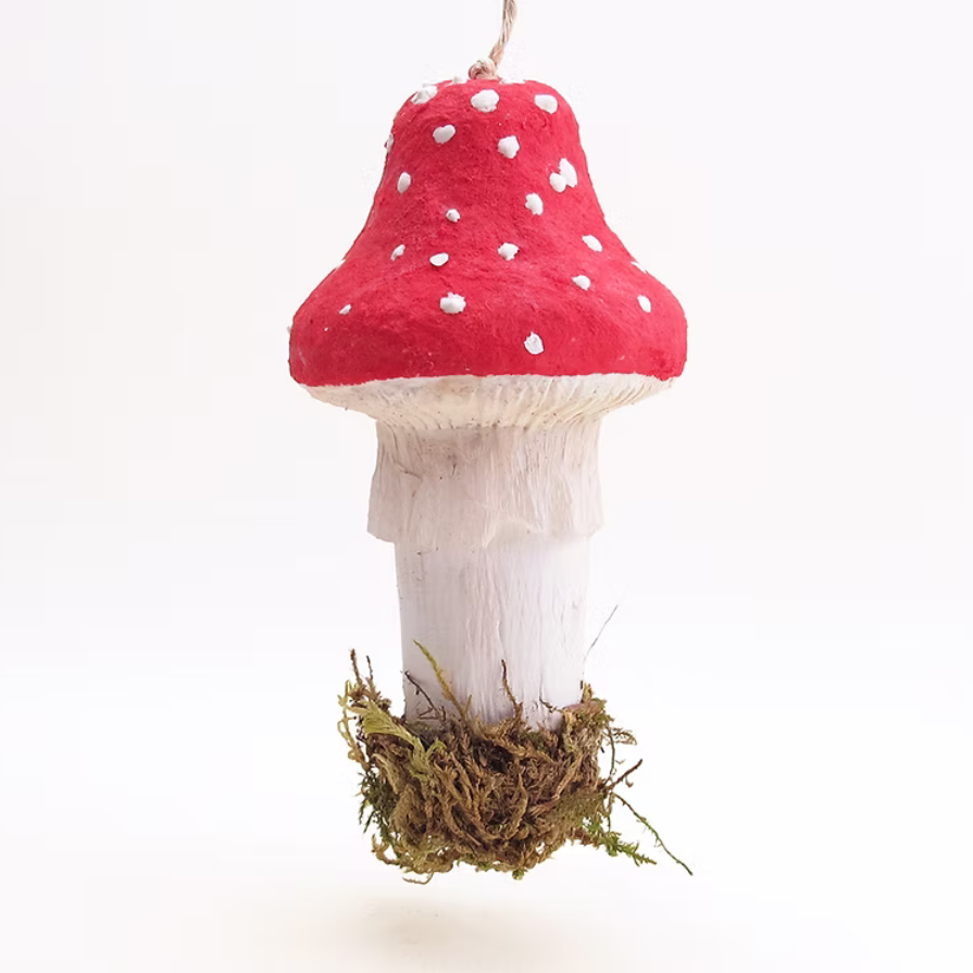 A small hanging mushroom ornament with a bright red cap, white spots, a white stem, and a tuft of brown moss at the base, suspended against a plain white background.