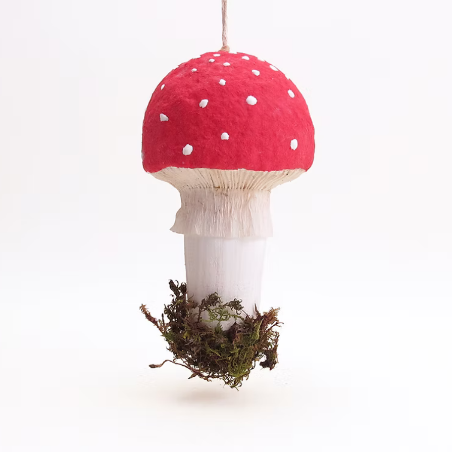 A hanging decorative mushroom ornament with a red felt cap, white spotted details, a white stem, and a small patch of moss at the base, shown against a plain white background.