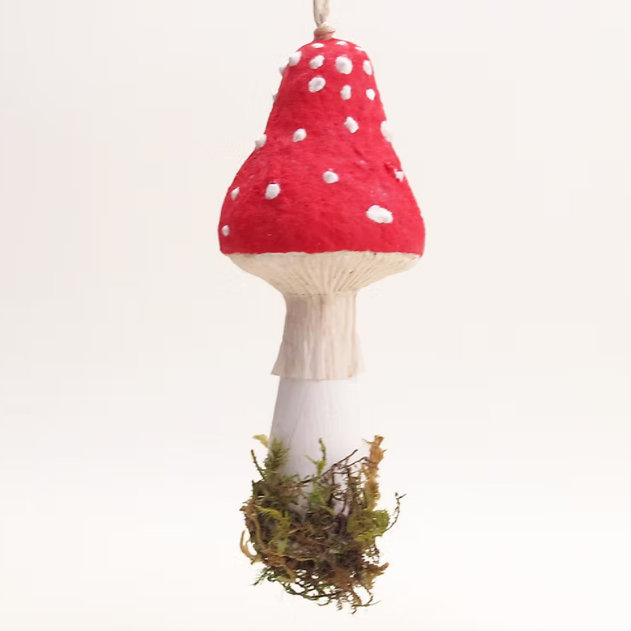 A felt mushroom ornament hanging against a plain white background. The ornament features a bright red felt cap with white spots, a white felt stem, and a base wrapped in green and brown faux moss.
