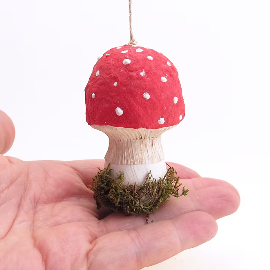 A small decorative mushroom ornament with a red cap, white spots, a light wood-toned stem, and green moss at the base, held in a person's hand against a white background.