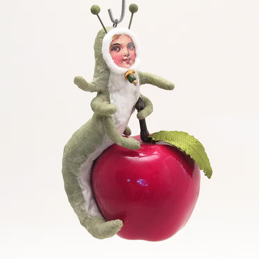 A whimsical hanging ornament featuring a green caterpillar-like creature wearing a white hood with a printed face, sitting on a glossy red apple with a green leaf, photographed against a plain white background.