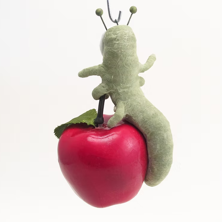 A small green felt caterpillar hanging from a metal hook, posed on top of a glossy red apple with a green leaf, against a plain white background.