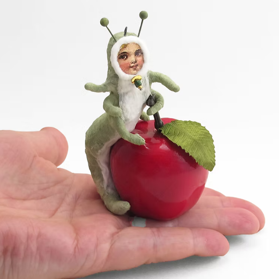 A small handmade figurine of a child in a green caterpillar-like costume sitting on a glossy red apple, displayed on an open human hand against a white background.