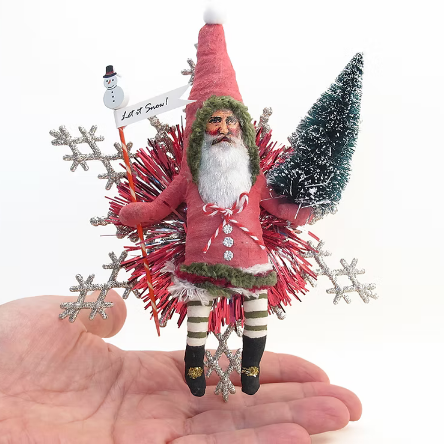 A hand holds a small festive Santa ornament against a white background. The ornament features a red hat and coat, white beard, striped legs, a small green tree, silver snowflakes, and red tinsel details, with a small sign reading 'Let It Snow!'.