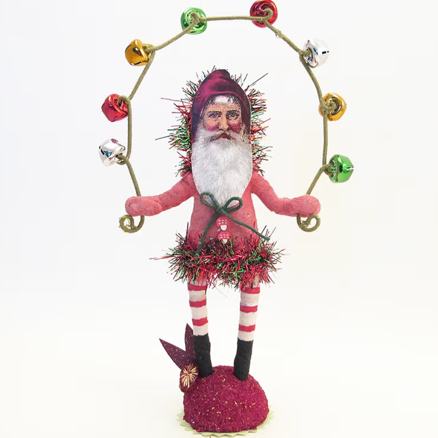 A festive Christmas nutcracker doll wearing a pink suit, white beard, and red hat, holding a gold hoop with colorful hanging bells, standing on a glittery magenta base against a white background.
