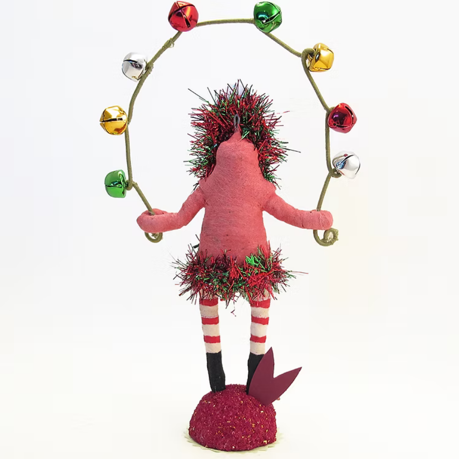 A handmade Christmas juggling doll with a pink body, red and white striped legs, and a green tinsel head and skirt, juggling a gold wire hoop decorated with colorful bells on a white background.