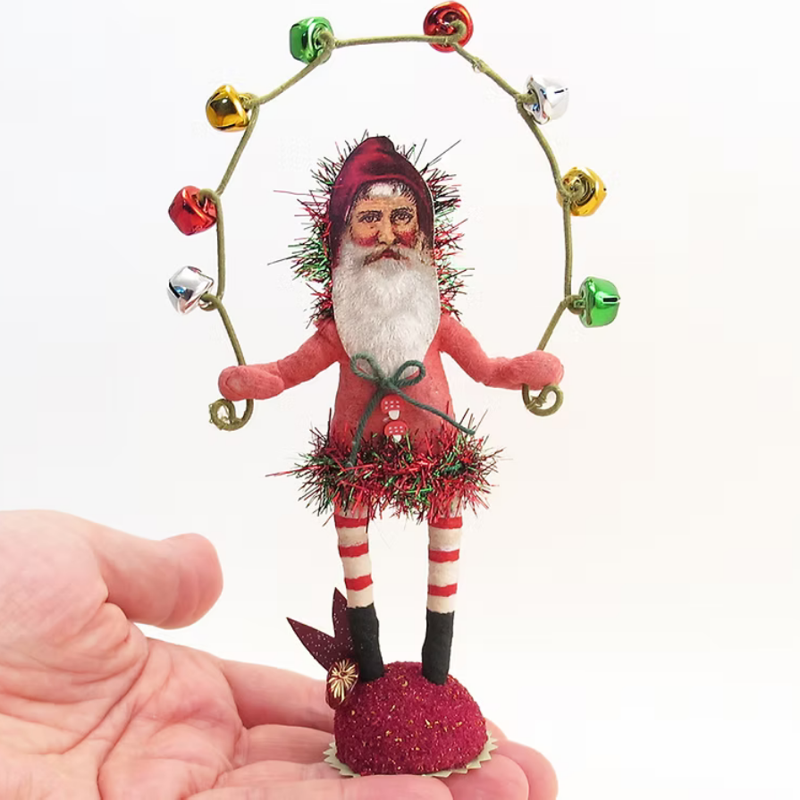 A small Santa Claus holiday ornament held in a person's hand against a plain white background. The ornament features a Santa figure with a white beard, red suit, and red-and-white striped legs, surrounded by a gold hoop with colorful bells.