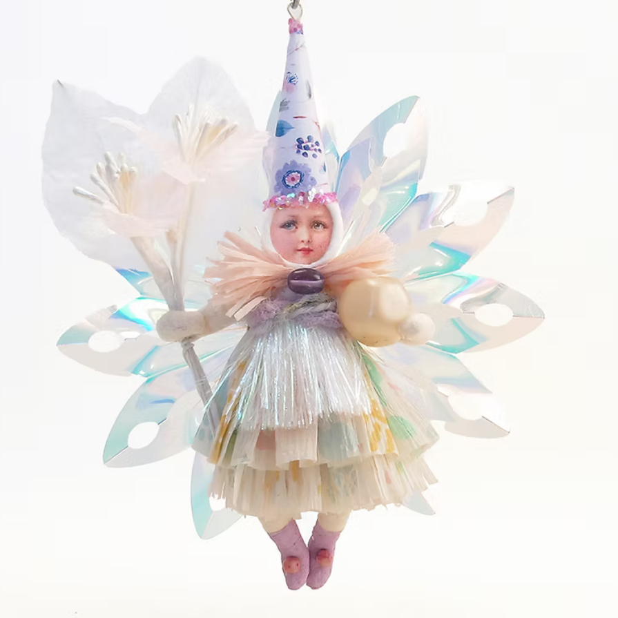 A whimsical fairy ornament hanging against a plain white background. The ornament features a small doll-like figure with a pointed lavender hat decorated with blue floral patterns, translucent iridescent wings in pale blue and white, a layered cream and iridescent dress, and pink shoes.