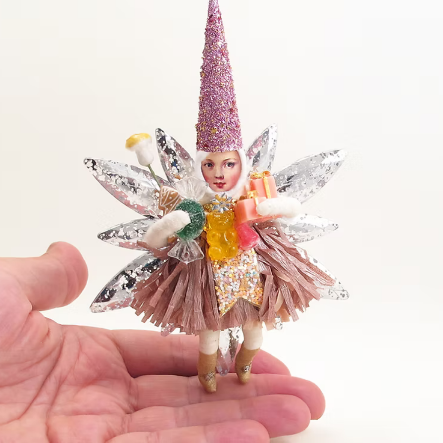 A miniature decorative fairy figurine with a glittery pink cone hat, silver wings, and a brown tulle skirt, displayed on a person's palm against a white background.