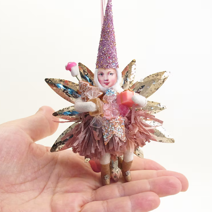 A miniature glitter fairy Christmas ornament held in a person's hand against a plain white background. The ornament features a pink glitter pointed hat, a pink glitter tattered skirt, gold glitter wings, and a small painted face.