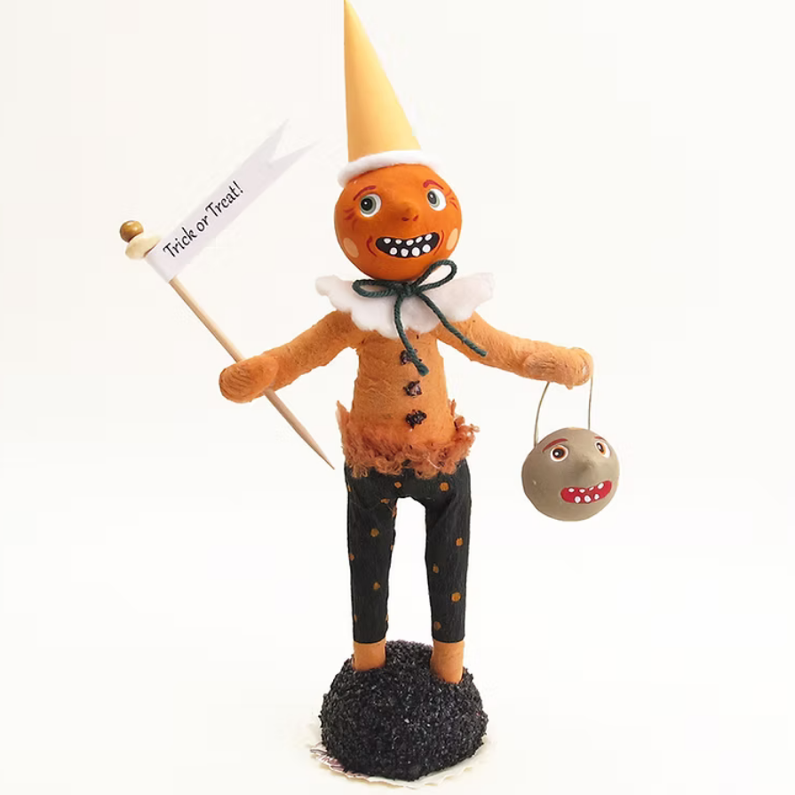 A handmade Halloween scarecrow figurine standing on a black textured base. The scarecrow has an orange face and body, a yellow cone hat, black pants with gold stars, and holds a 'Trick or Treat!' flag and a gray monster bucket.