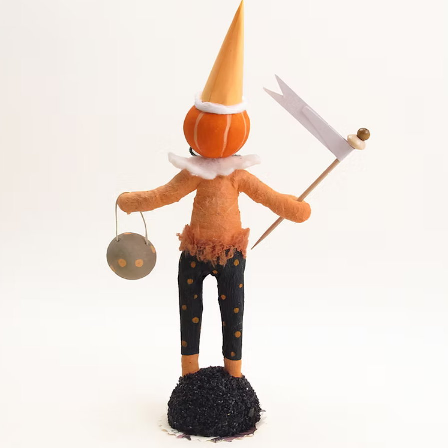A handmade orange pumpkin doll figurine with a tall orange cone hat, white ruffled collar, black pants with gold polka dots, holding a small gray lantern and a white flag, standing on a black base against a white background.