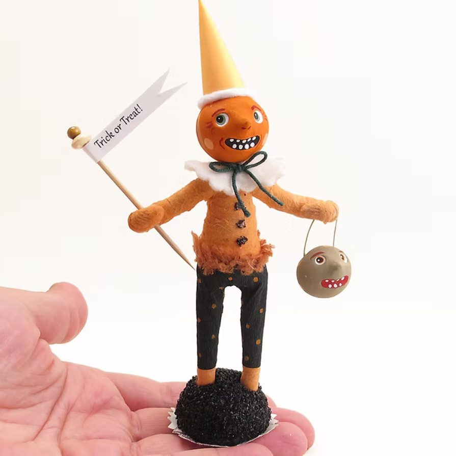 A small orange Halloween scarecrow figurine with a yellow cone hat, white ruffled collar, black bow, black pants with orange stars, and a sign reading 'Trick or Treat', displayed on a person's hand against a white background.