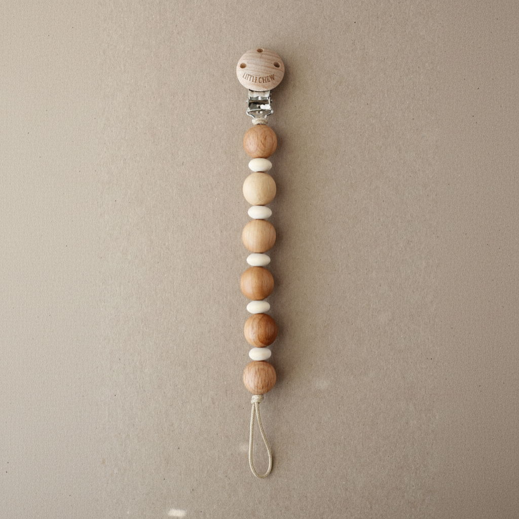 A wooden bead pacifier clip with natural wood and white beads, featuring a round wooden top cap with a smiley face and a silver metal clasp, displayed on a textured beige background.