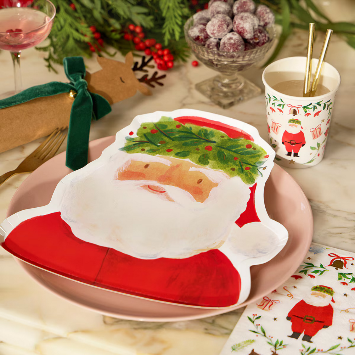A festive Christmas table setting featuring a large Santa Claus-shaped paper plate on a pink charger plate, with matching Santa-themed napkins, a paper cup with gold straws, and holiday decorations in the background.