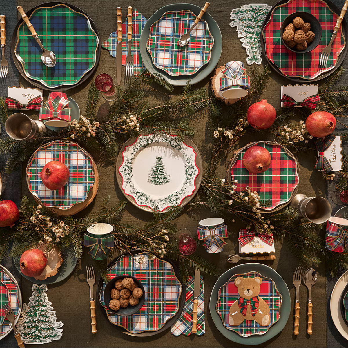 A festive holiday table setting featuring coordinated Christmas-themed tableware. The place settings include plaid napkins, green-rimmed plates with plaid and tree motifs, a central white plate with a green tree design, and matching cups and bowls.