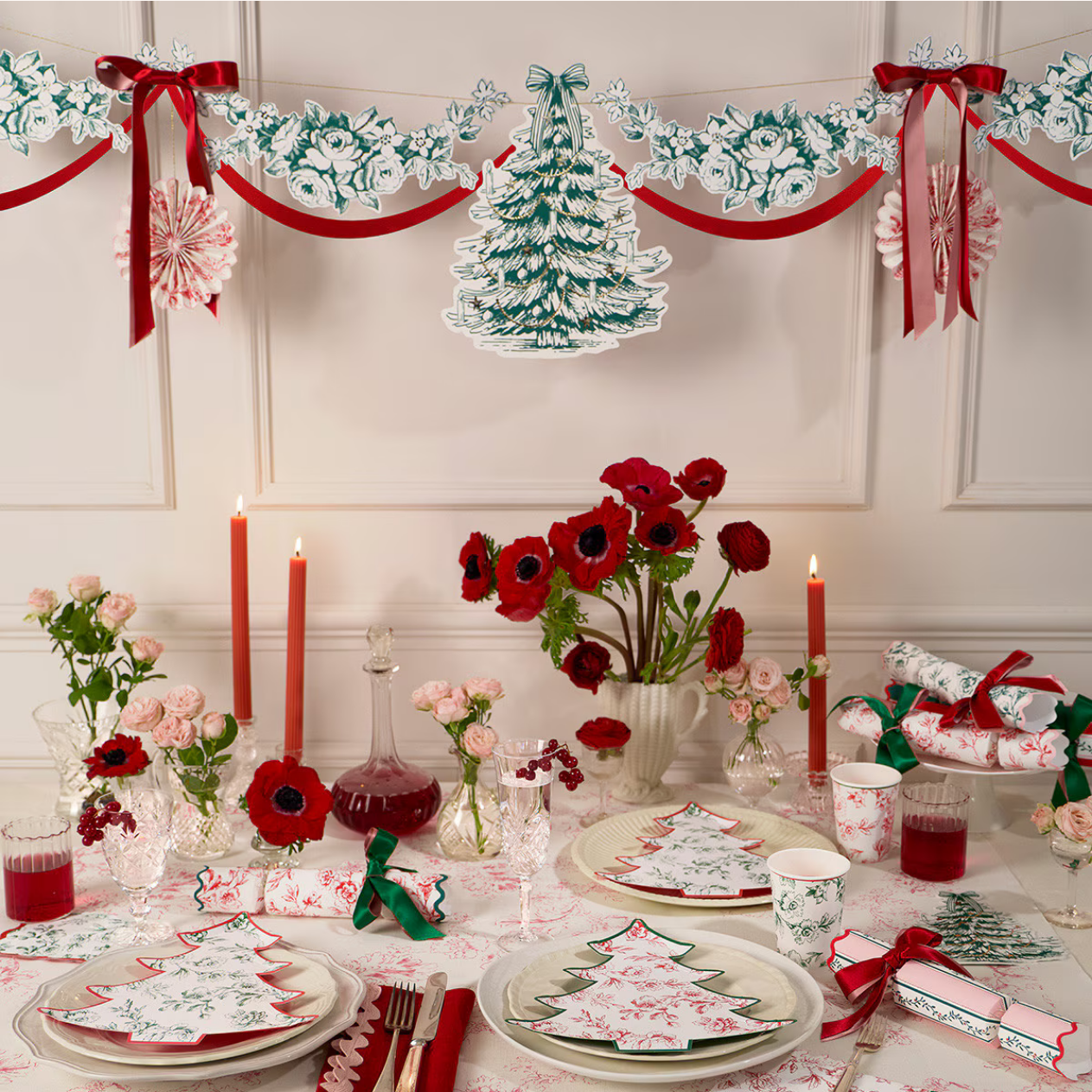 A festive Christmas table setting decorated with red and white table linens featuring tree patterns, red candles, red poppy flowers, and a matching red and white garland with a central tree cutout against a white paneled wall.