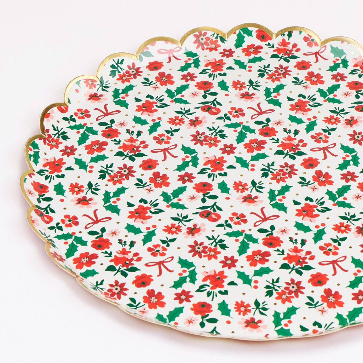 A scalloped ceramic serving platter with a festive Christmas pattern of red holly leaves, red bows, and green berries on a white background, finished with a gold rim.