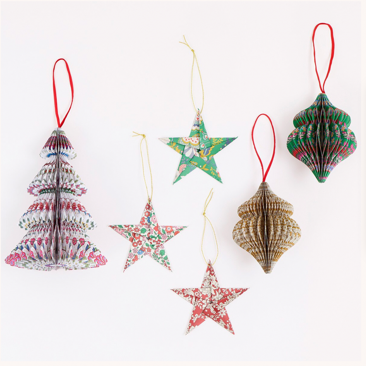 Six decorative hanging ornaments made from folded patterned paper are arranged on a plain white background. The set includes Christmas tree, star, and shell-shaped ornaments in assorted multicolor floral and geometric patterns, each with a red, gold, or yellow hanging loop.