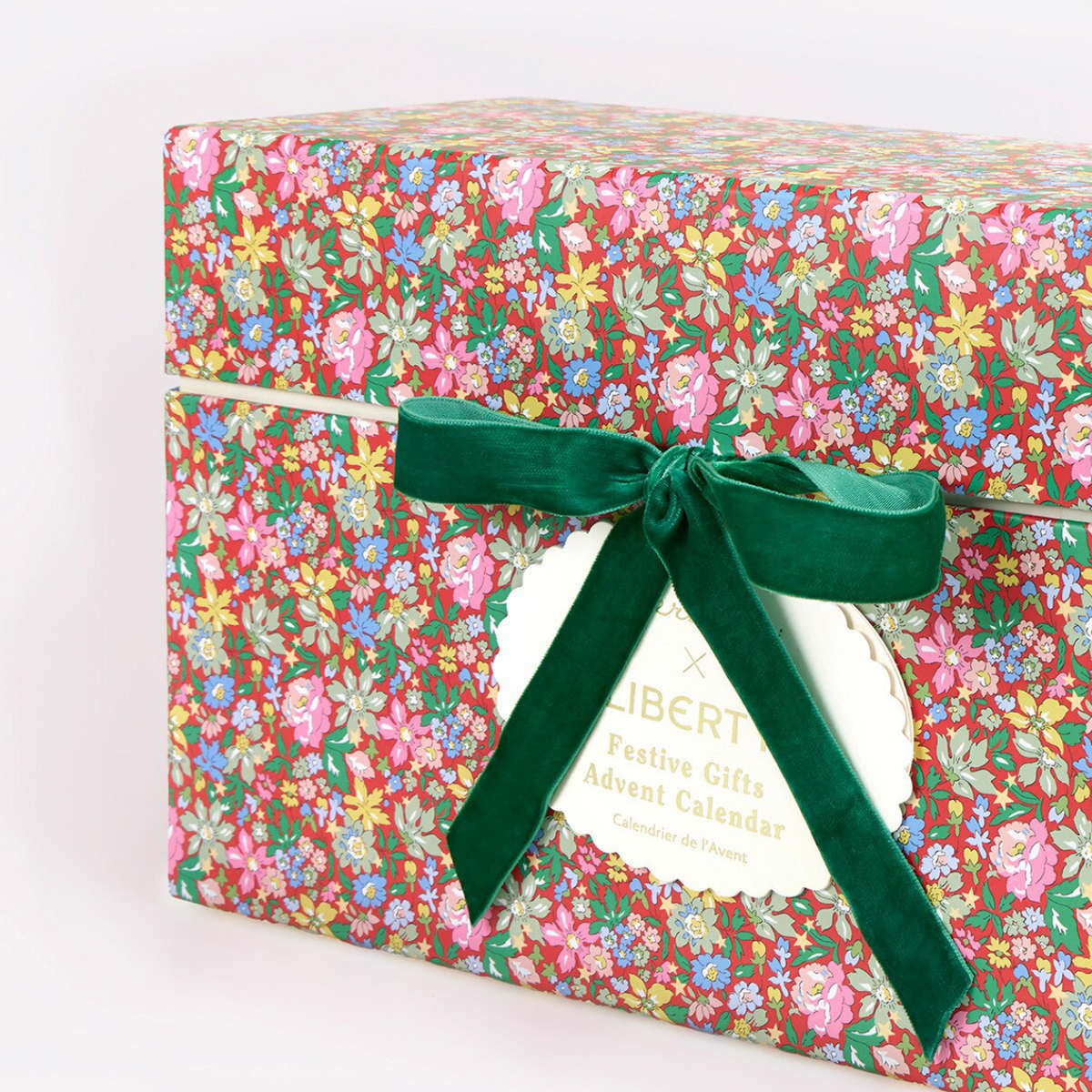 A square gift box wrapped in a colorful floral patterned paper with pink, yellow, blue, and green flowers on a dark background, tied with a dark green velvet ribbon and bow. A white paper tag is attached and reads 'LIBERTY Festive Gifts Advent Calendar'.