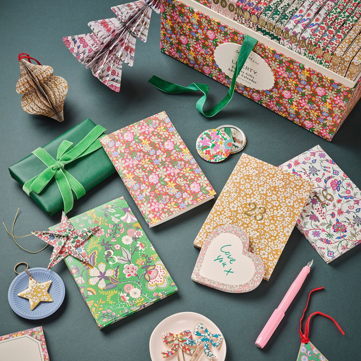 A styled flat-lay of multiple floral and patterned gift boxes, notebooks, paper ornaments, a green ribbon-wrapped package, small accessories, and a pink pen arranged on a dark teal background.