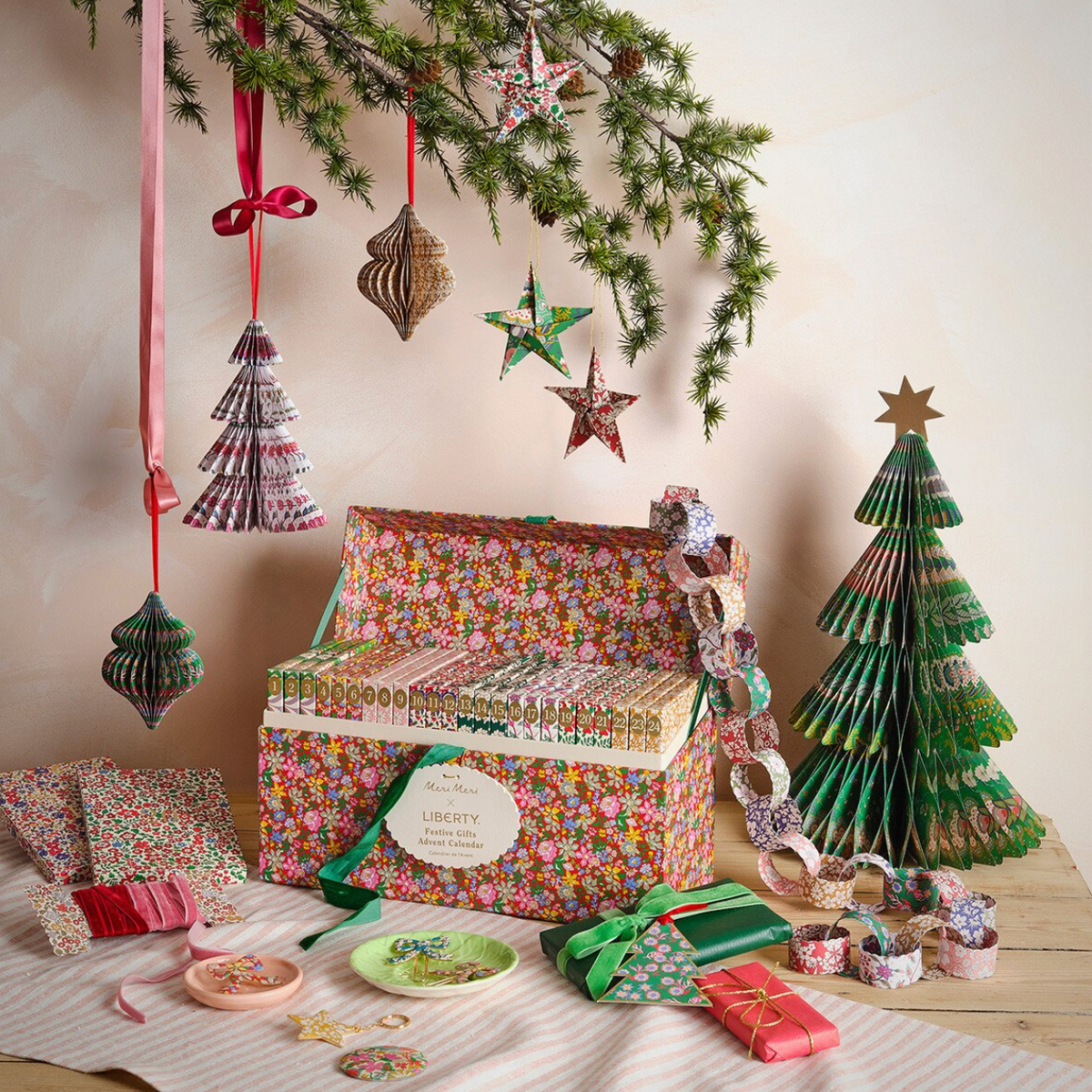 A styled holiday gift set featuring a floral paper gift box labeled 'Liberty Paper Company', matching patterned gift bags, colorful paper washi tapes, small wrapped presents, and decorative hanging ornaments under a pine branch.