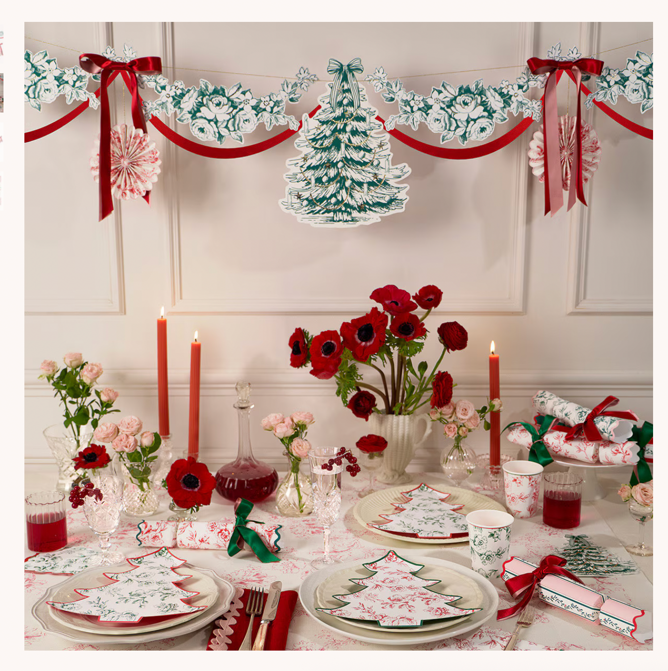 A festive Christmas table setting decorated with red and white table linens, red candles, red poppy flowers, wrapped crackers, and a matching hanging garland with a tree silhouette against a paneled wall.