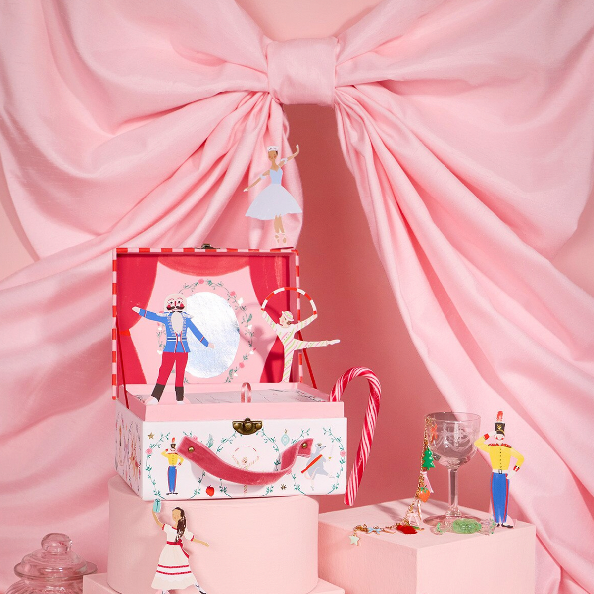 A pink-themed decorative music box display with a large pink bow backdrop. The main product is a tiered pink storage box set with a red-lidded music box on top, featuring illustrated figures, candy canes, and small figurines arranged around it on pink pedestals.