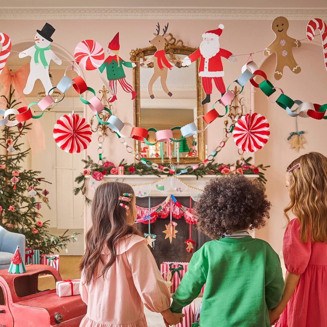 Three children standing in front of a decorated Christmas fireplace, looking up at a festive garland with Santa, snowman, reindeer, gingerbread, candy canes, and paper chains hanging above them in a warm, decorated living room.