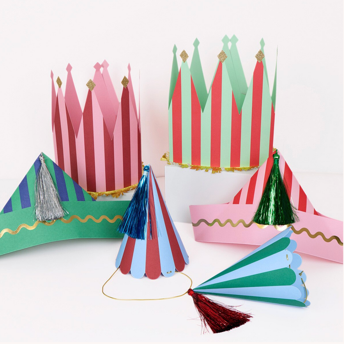 A set of colorful paper party hats and crowns displayed on a white surface against a white background. The hats include striped designs in pink, red, green, blue, and purple with tassel accents, and one crown features gold glitter trim.