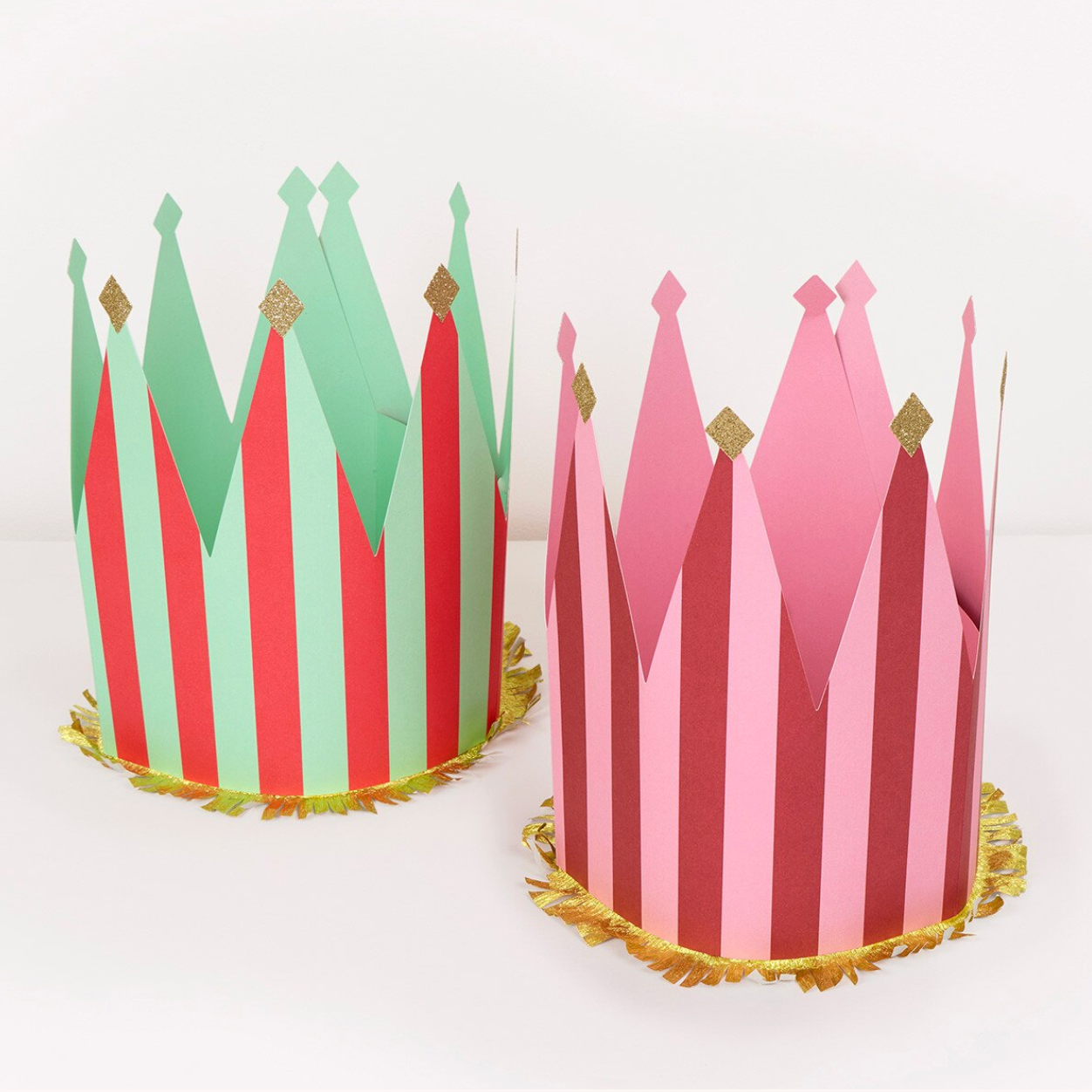 Two paper crown party hats on a white background, one in green and red stripes with gold accents, and the other in pink and red stripes with gold accents.