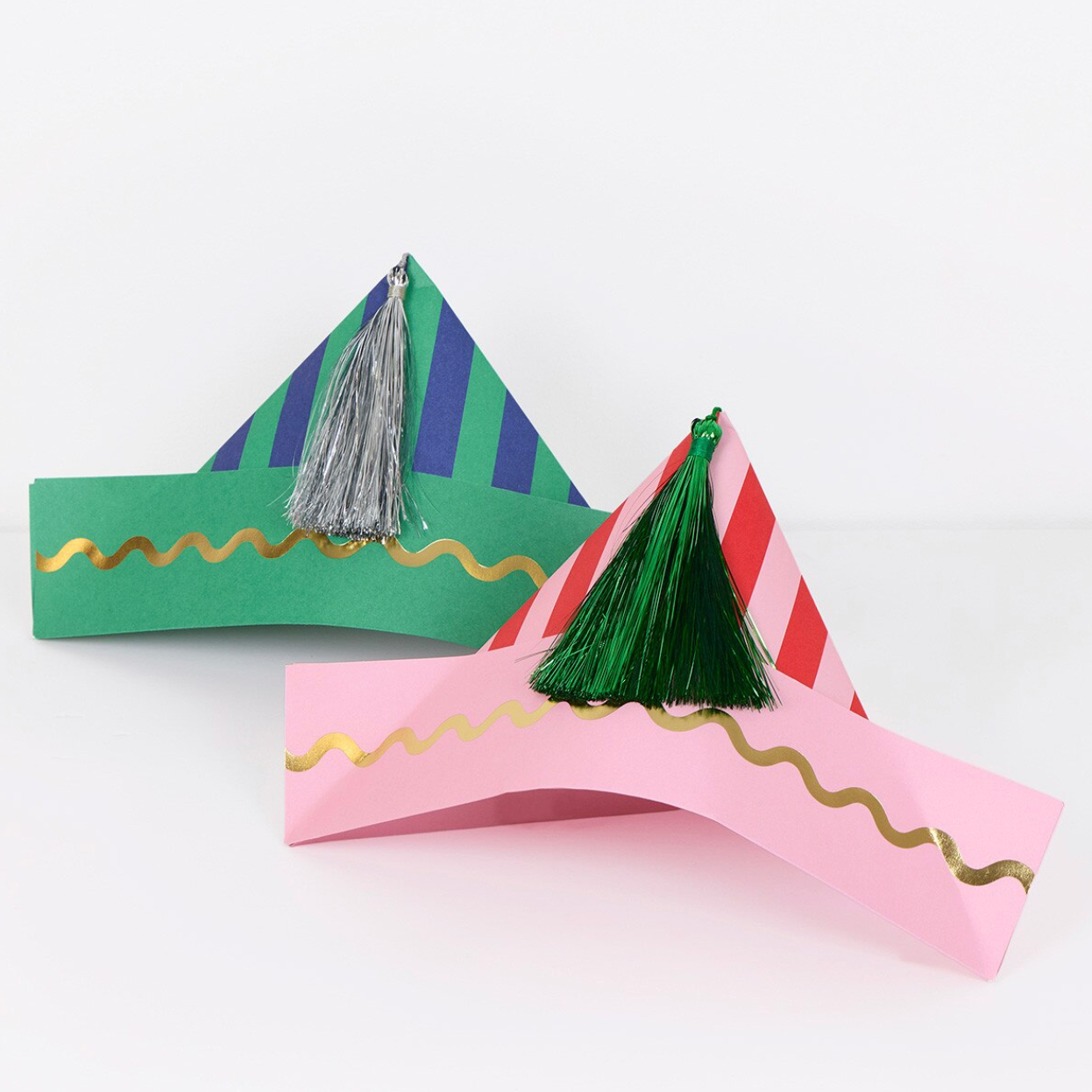 Two handmade paper party hats on a white background. One hat is green with blue and purple diagonal stripes, a silver tassel, and gold foil wavy trim.
