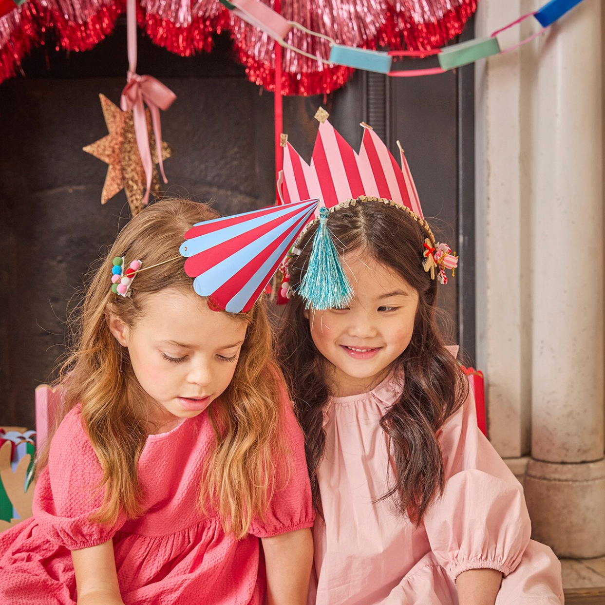Two young girls wearing red and white striped party hats with blue tassels, seated together in a decorated indoor setting with festive garlands and a star hanging in the background.