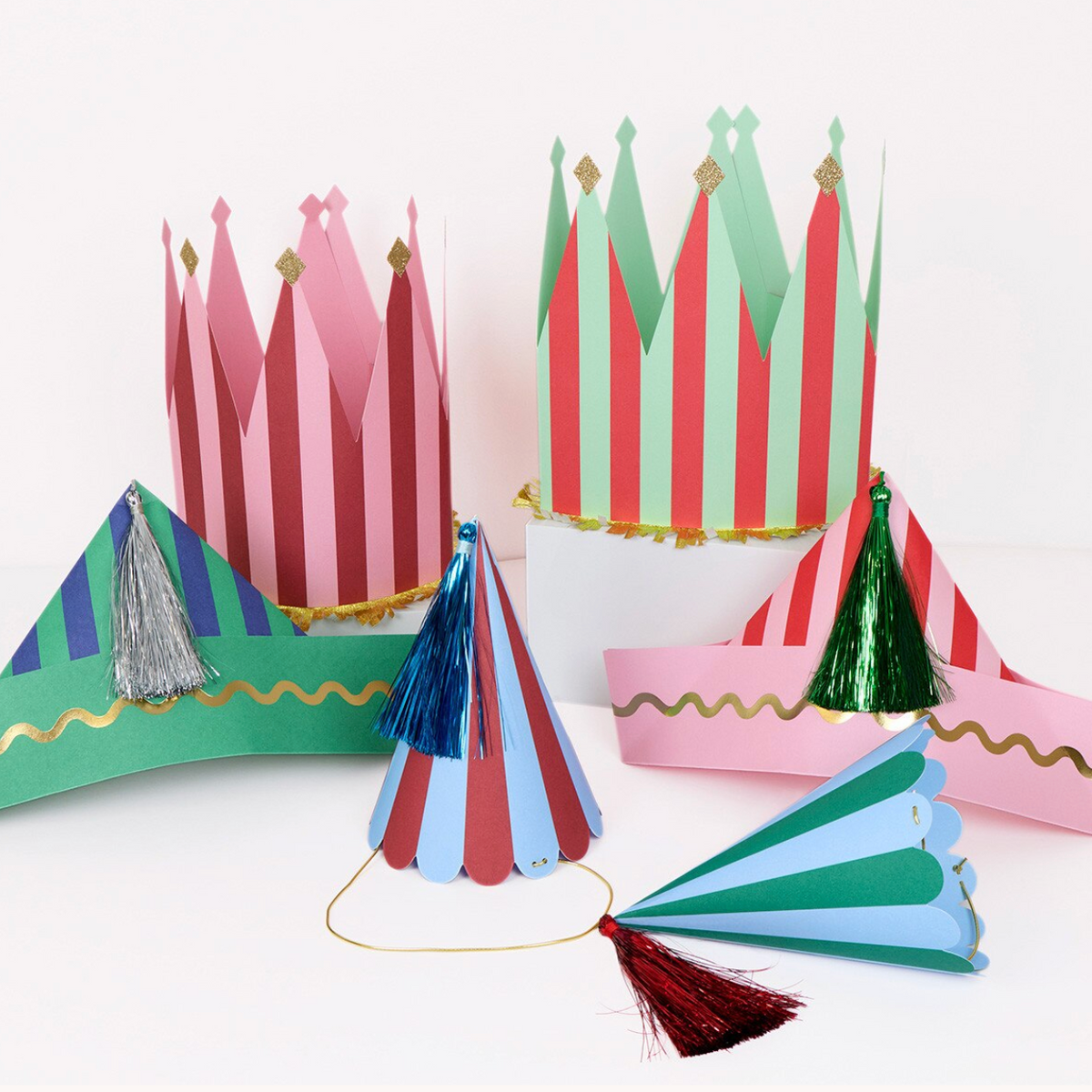 Colorful paper party decorations including two tall pointed crowns, a curved banner, and two cone hats, arranged on a white background.
