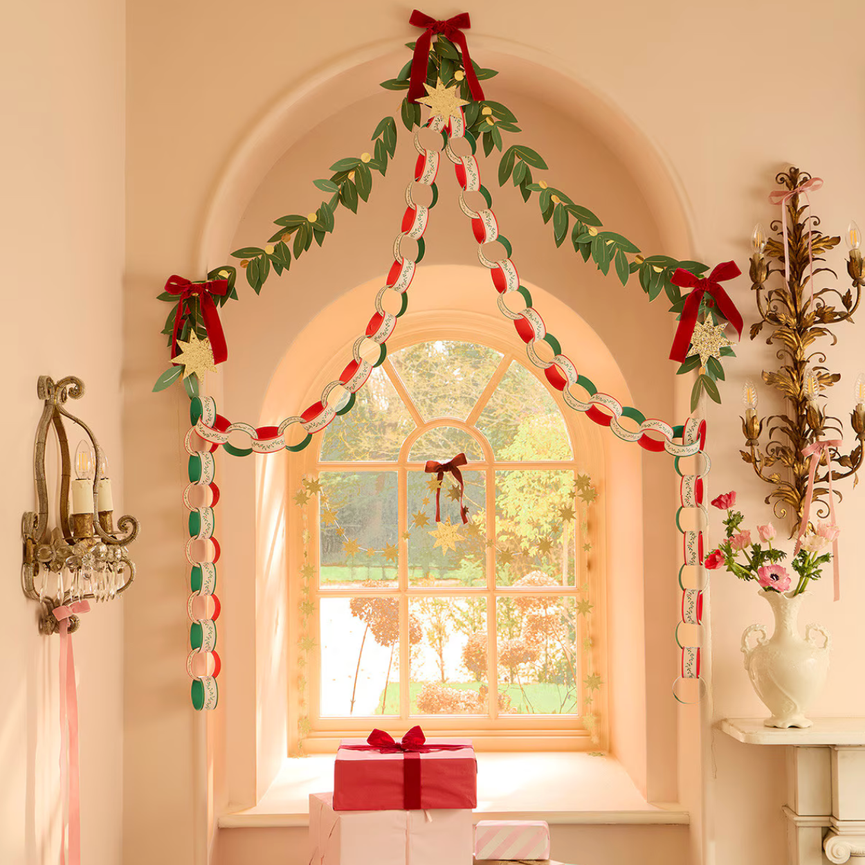 A festive holiday garland decoration displayed in an arched window frame on a peach-colored wall. The garland features green leafy vines, red bows with white pom-pom tails, and white star ornaments, with a matching red and white gift box placed below the window.