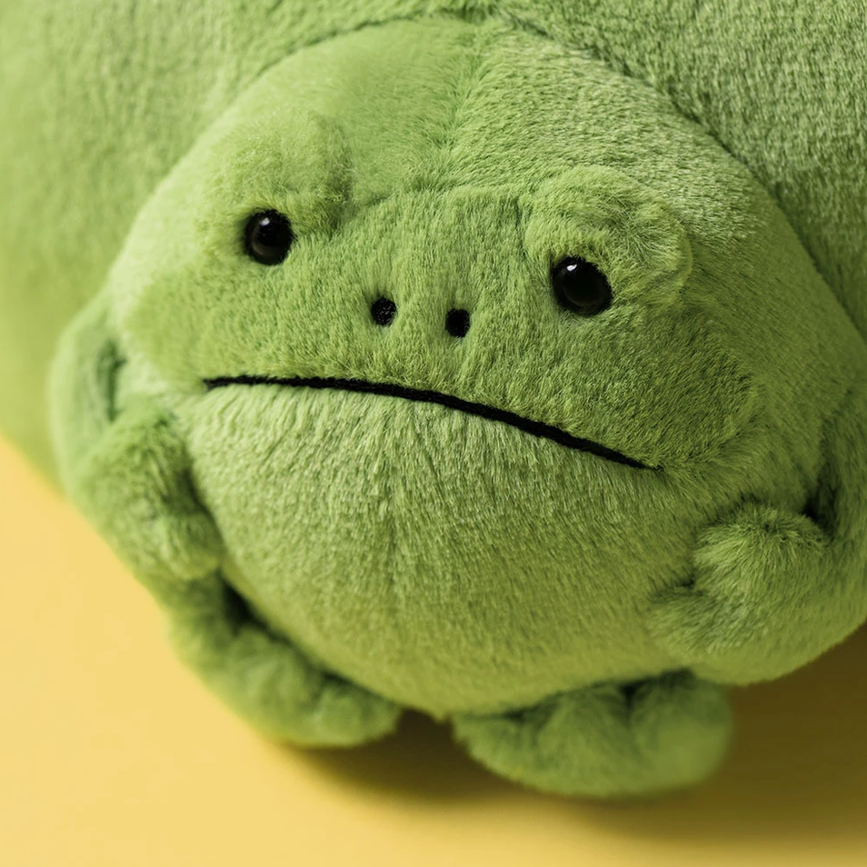 A green plush frog toy with a sad, downturned expression, photographed close up against a soft yellow background.