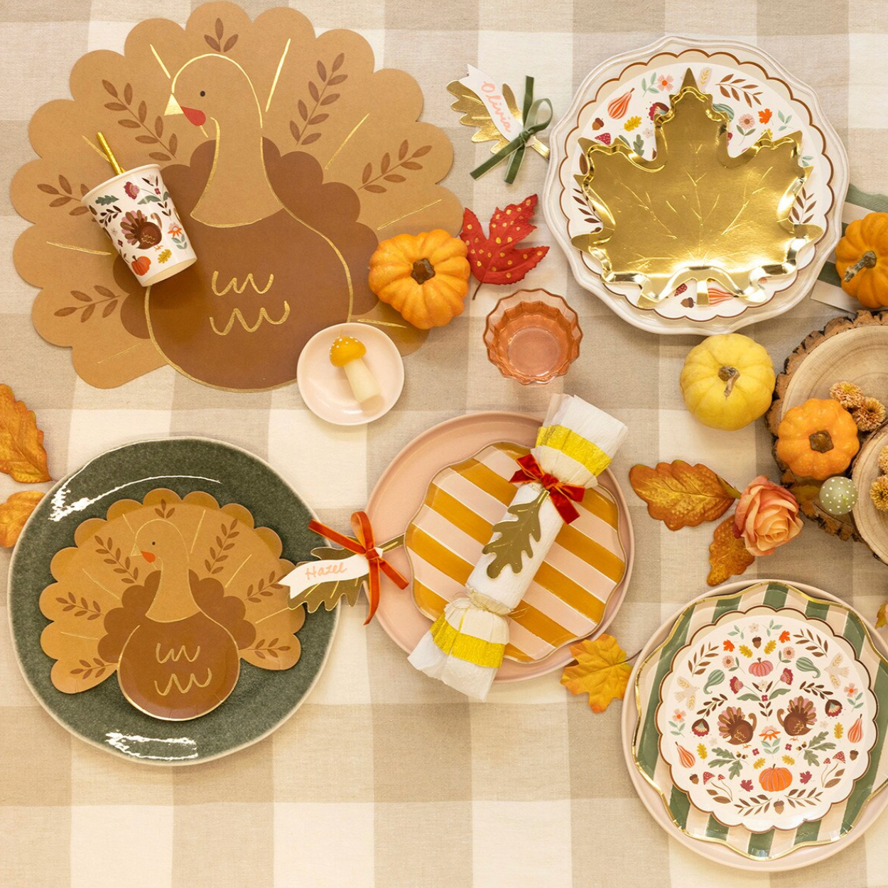 A styled Thanksgiving table setting with autumn-themed ceramic plates, napkins, placemats, and small pumpkins arranged on a beige checkered tablecloth.