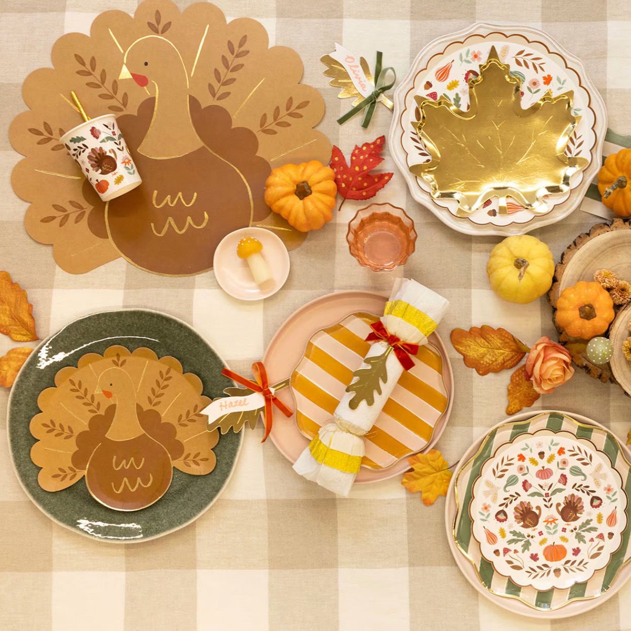 A styled Thanksgiving table setting with coordinated autumn tableware and decor on a beige-and-white checkered tablecloth. Visible items include layered plates with floral and gold leaf designs, a large turkey-shaped placemat, napkins wrapped in striped paper with ribbon ties, small pumpkins, fall leaves, and a cup with a straw.