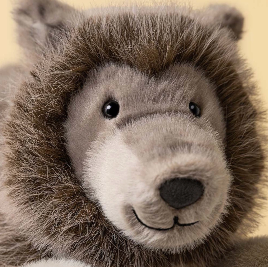 Close-up of a plush stuffed animal head with brown and gray fur, black eyes, and a dark nose against a pale yellow background.