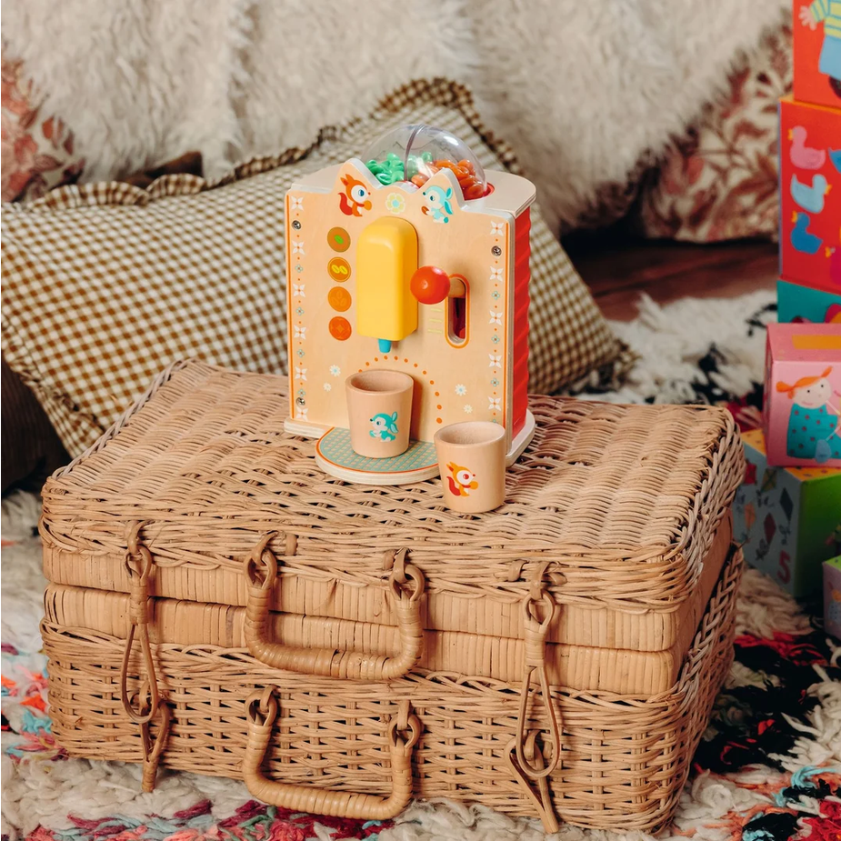 A children's toy play set featuring a small multicolor juice bar machine and cups placed on top of a woven wicker picnic suitcase, styled in a cozy living room setting.