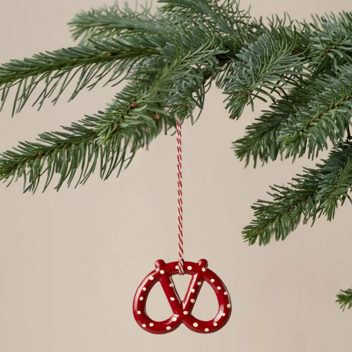 A red pretzel-shaped Christmas ornament with white polka dots hangs from a red-and-white striped string on a green pine tree branch against a plain beige background.