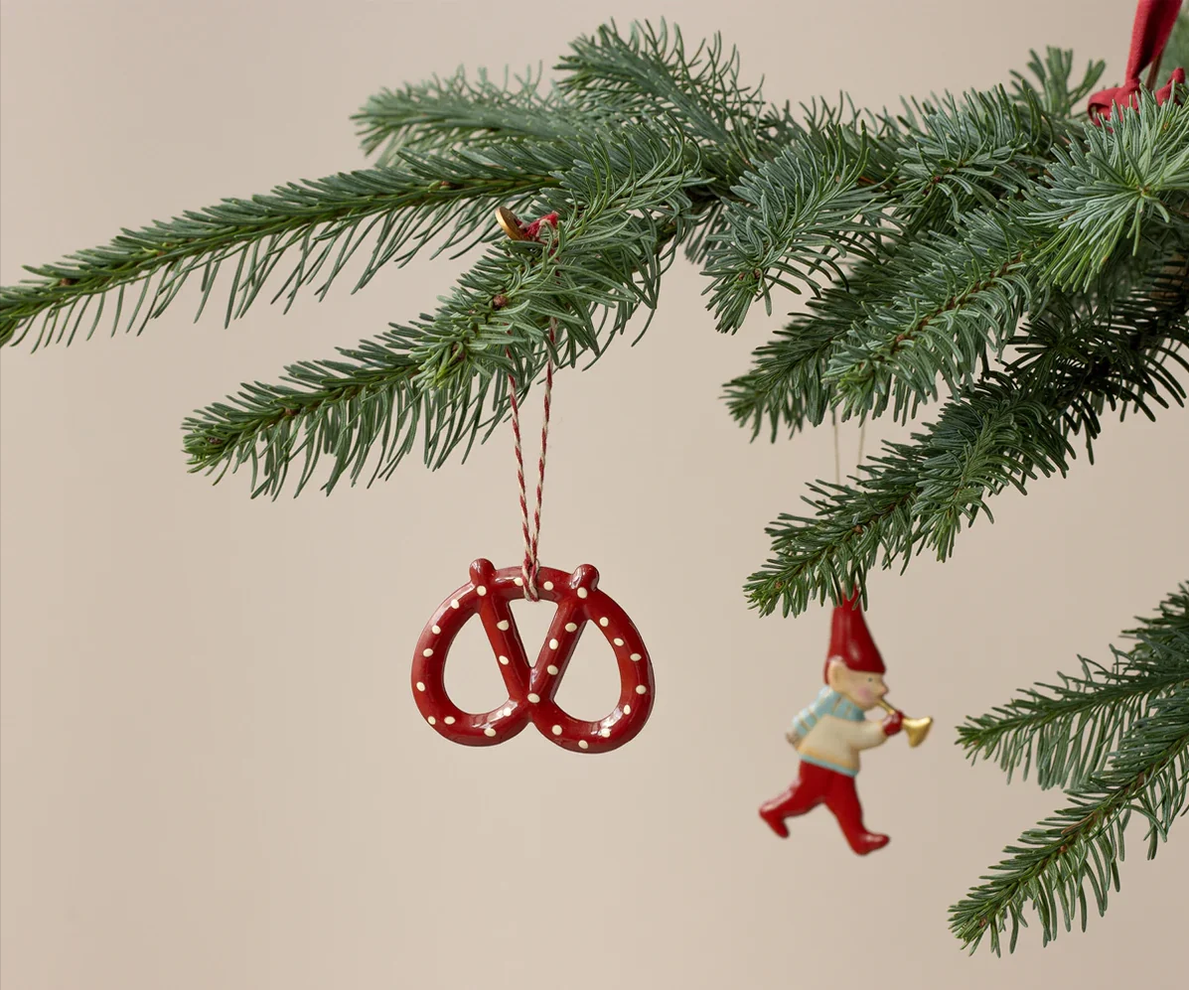 A red pretzel-shaped Christmas ornament with white polka dots hangs from a green evergreen tree branch against a plain beige background, with a small gnome ornament nearby.