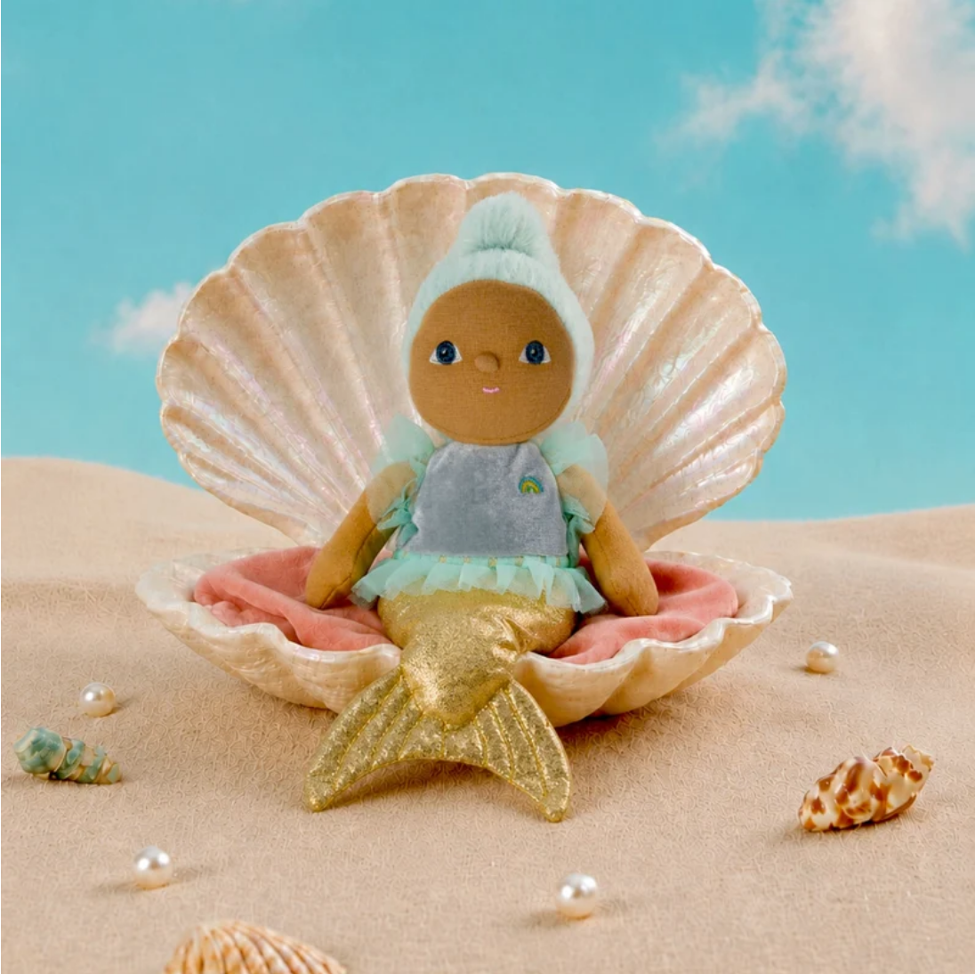 A small mermaid doll with brown skin, blue eyes, mint hair, and a gold tail sits inside a large beige seashell on pink sand, with a blue sky backdrop and scattered shells around.