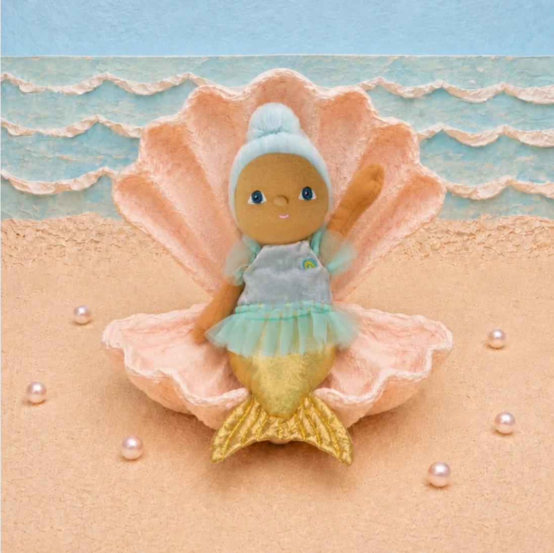 A mermaid doll with  blue hair and tutu sits inside a large peach seashell on a sandy beach backdrop with blue ocean waves behind it.