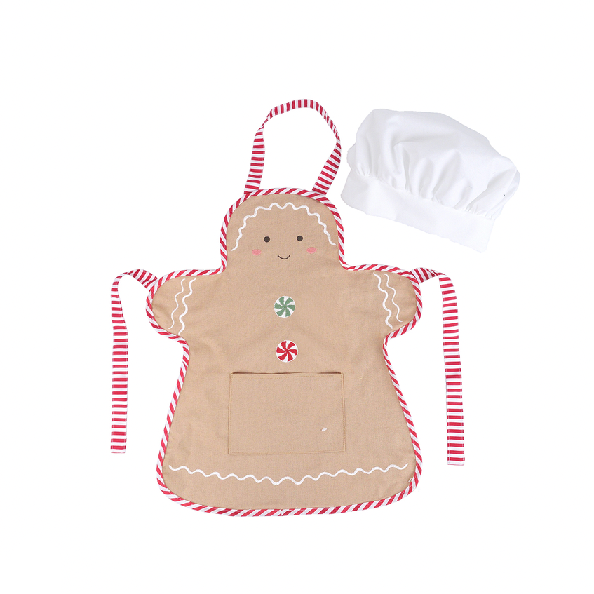 A gingerbread-themed fabric apron in beige with red and white striped straps and trim, decorated with embroidered candy details, shown alongside a white chef's hat on a plain white background.