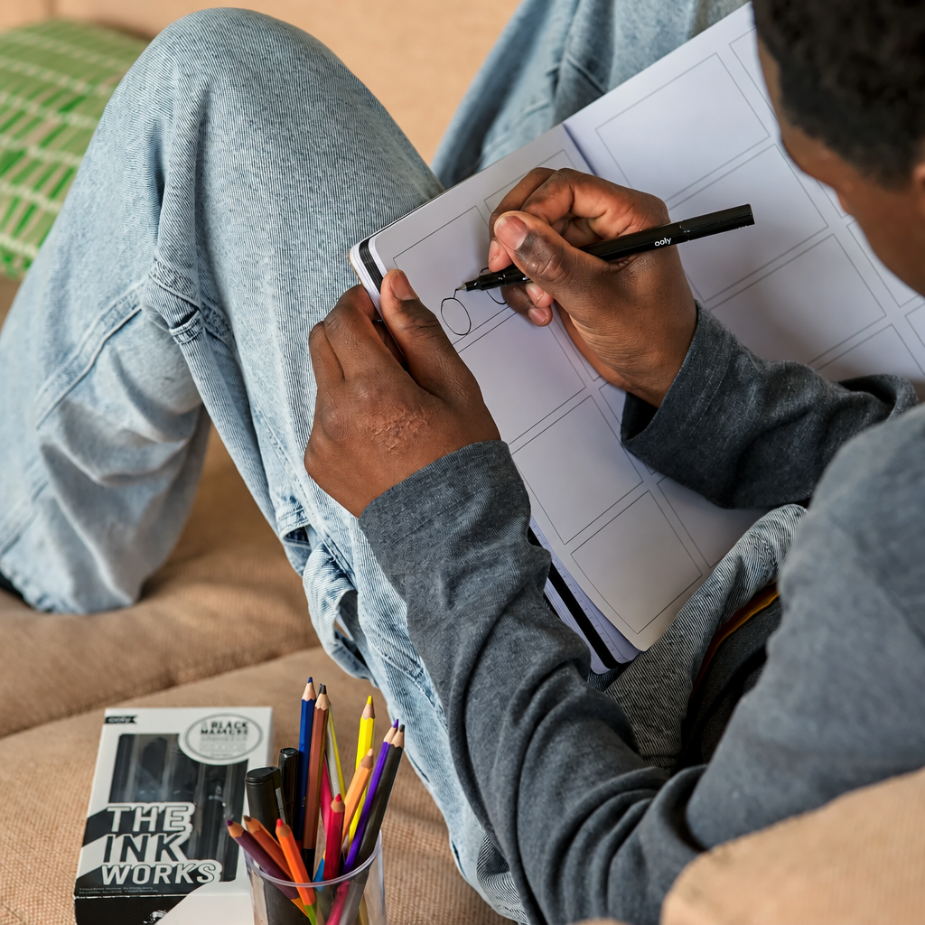 A person sitting on a beige couch draws in a notebook with a black pen. A box of The Ink Works black pens and a cup of colored pencils are visible on the couch beside them.