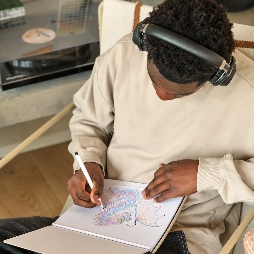 A person wearing beige sweatpants and black over-ear headphones sits in a chair, drawing a colorful dragon in a sketchbook with a white pen.