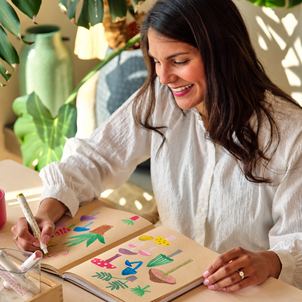 A woman with long dark hair, wearing a white blouse, smiles while drawing in a notebook with colorful mushroom illustrations. She is seated in a bright, sunlit room with green plants in the background.