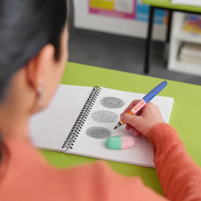 A person in an orange sweater sits at a green desk, writing in a spiral notebook with a blue and orange pen. On the notebook are three gray spiral drawings, and next to the pen is a pastel eraser.
