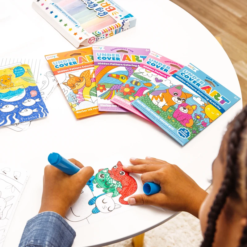 Child coloring with markers on a table with 'Cover Art' coloring books.